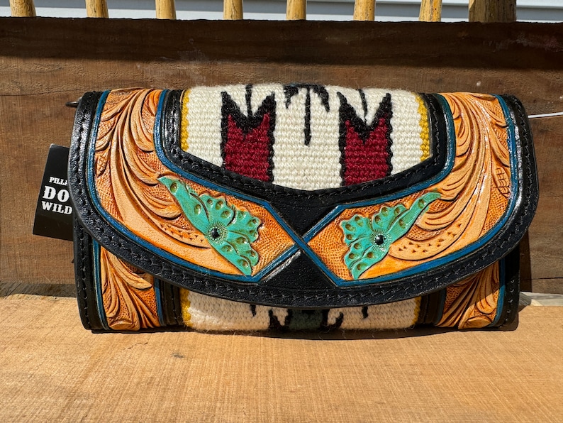 May include: A brown leather wallet with tooled floral designs and turquoise accents. The front flap features a woven panel with red and white stripes and the text "MTM".