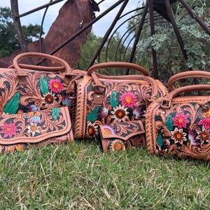 May include: A collection of three tan leather handbags and two matching wallets featuring intricate floral tooling with hand-painted accents in pink, yellow, and green. Each item displays a dark background within the carved leather patterns.