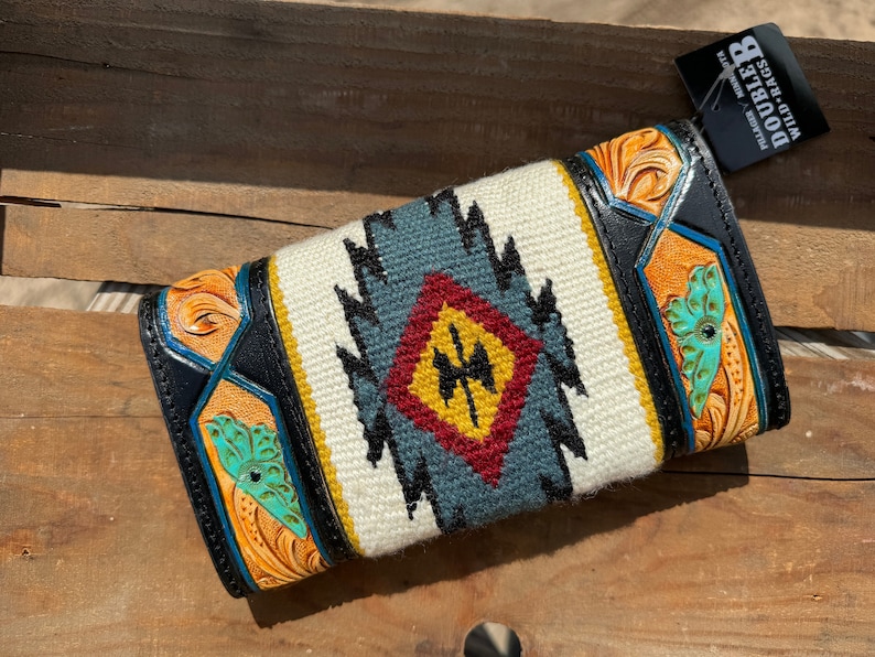 May include: A black leather wallet with a white, blue, red, and yellow geometric pattern. The wallet has a tooled leather design with a butterfly motif. The brand name "WILD HAGS DOUBLE B" is visible on a tag.