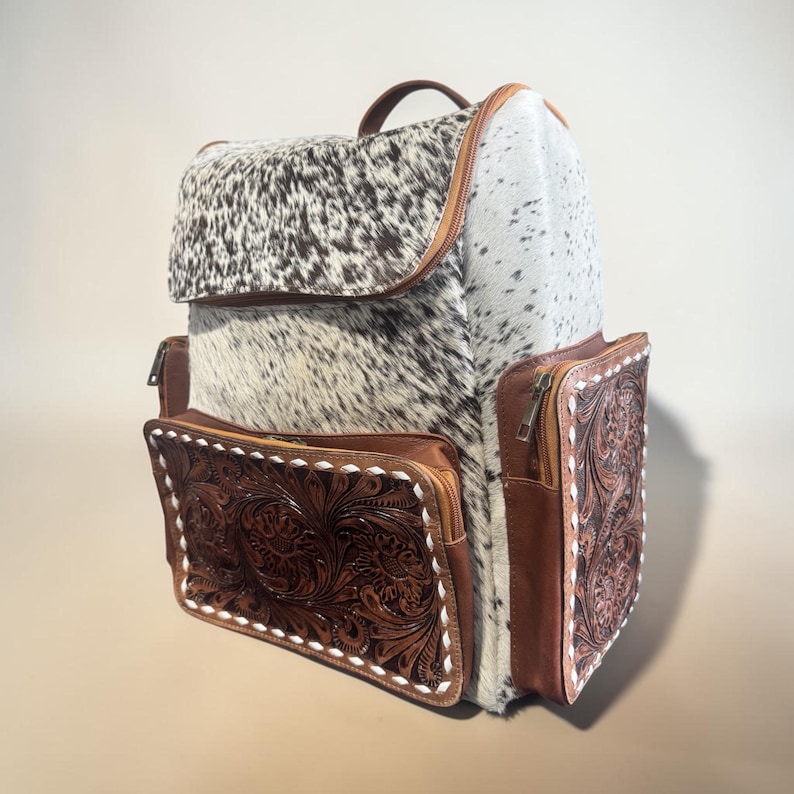 May include: A western-style backpack with a cowhide flap and sides. The backpack features brown leather pockets with floral tooling and white stitching. The zipper pulls and handle are brown leather.