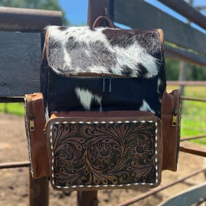 May include: A cowhide backpack with a black and white pattern on the flap. The front pocket is brown leather with a floral tooled design and white stitching. The sides have brown leather pockets with zippers. The bag has a top handle and adjustable straps.