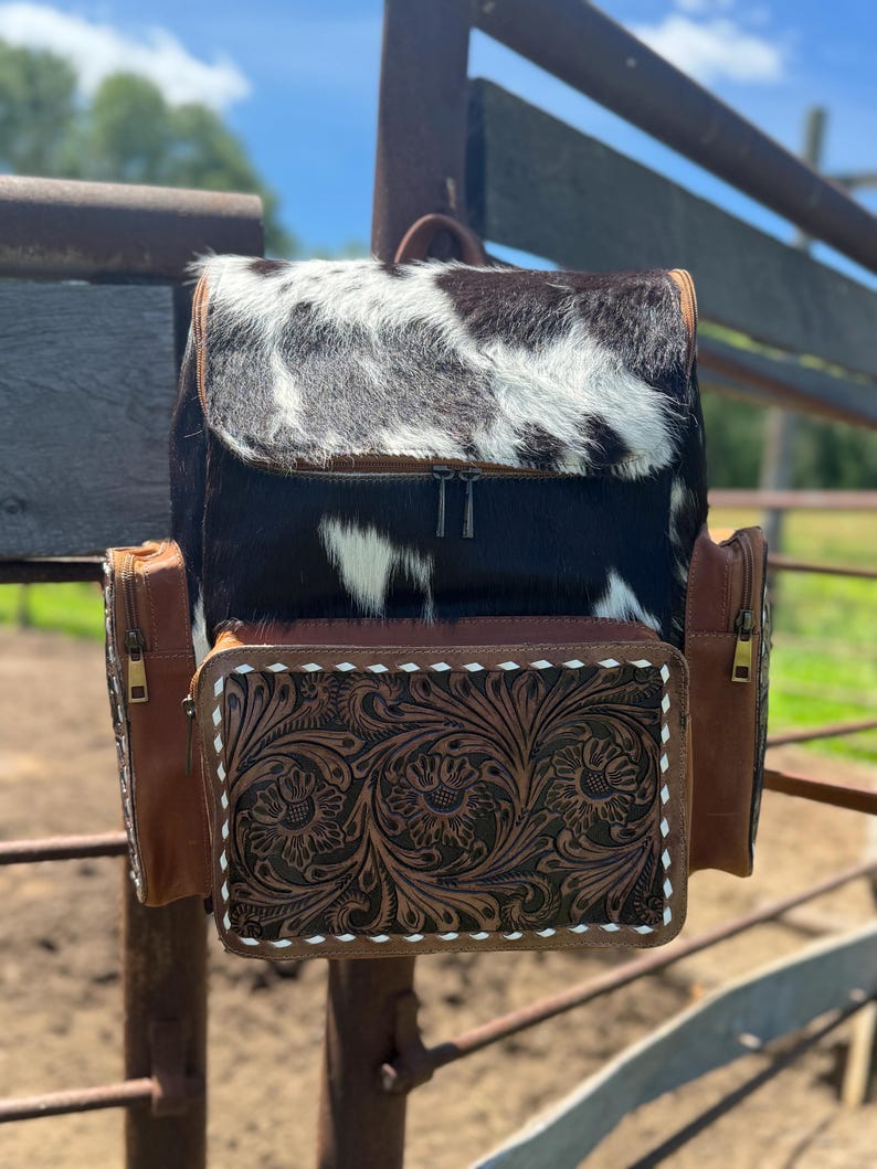 May include: A cowhide backpack with a black and white pattern on the flap. The front pocket is brown leather with a floral tooled design and white stitching. The sides have brown leather pockets with zippers. The bag has a top handle and adjustable straps.