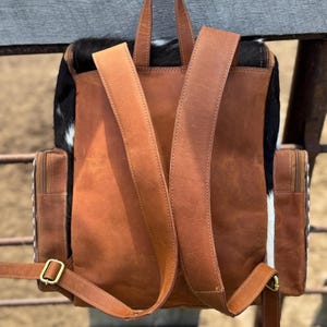 May include: A brown leather backpack with adjustable straps and a top handle. The backpack features a black and white cowhide panel on the top and sides, with two side pockets and gold-colored hardware. The bag is designed for everyday use.