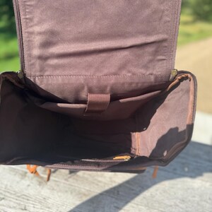 May include: Open view of a brown leather backpack with multiple interior pockets. The bag has a rectangular shape with a flap that opens to reveal the interior. The interior is lined with a lighter brown fabric. The bag has a handle and straps.