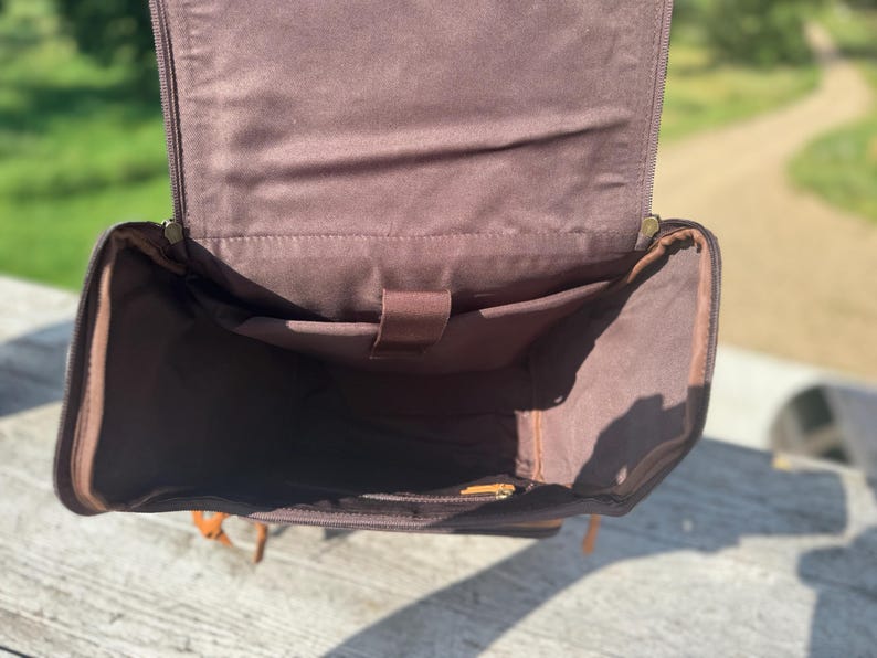 May include: Open view of a brown leather backpack with multiple interior pockets. The bag has a rectangular shape with a flap that opens to reveal the interior. The interior is lined with a lighter brown fabric. The bag has a handle and straps.