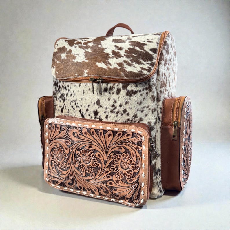 May include: A cowhide backpack with brown and white spotted pattern, featuring brown leather trim and accents. A matching brown leather wallet with floral tooling is in front. The backpack has two side pockets and a top handle.