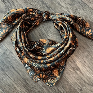 Puede incluir: Un pañuelo negro y naranja con estampado de cachemira, anudado en la parte superior. El pañuelo está hecho de un material sedoso y está drapeado sobre una superficie de madera. El diseño de cachemira presenta detalles en naranja y blanco.