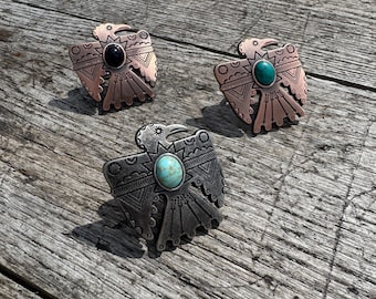 May include: Three silver and copper colored metal bird pendants with turquoise and black stones. The pendants are on a wooden surface.