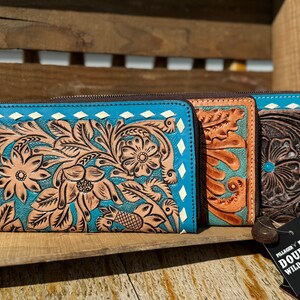 May include: Three leather wallets with floral tooling. The wallets are in shades of turquoise, tan, and brown. Each wallet has a zipper closure and a decorative diamond pattern around the edges. The wallets are displayed on a wooden surface.
