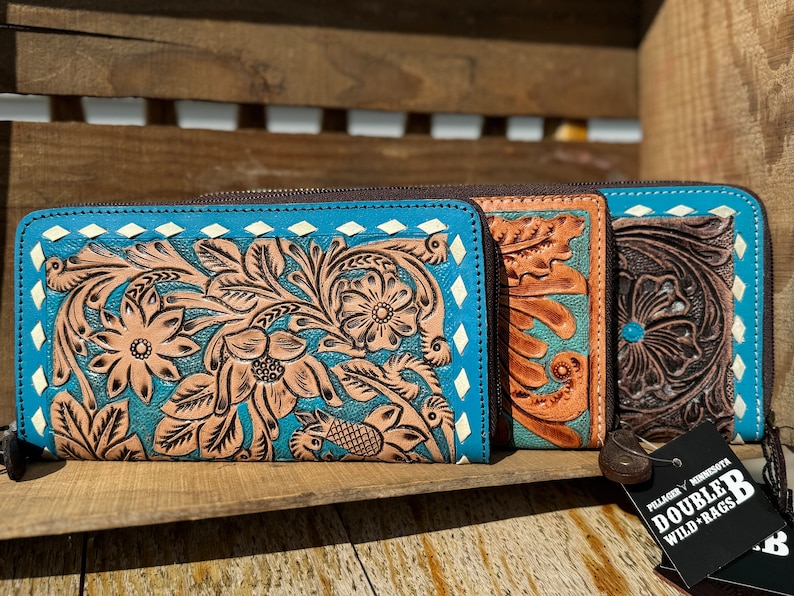 May include: Three leather wallets with floral tooling. The wallets are in shades of turquoise, tan, and brown. Each wallet has a zipper closure and a decorative diamond pattern around the edges. The wallets are displayed on a wooden surface.