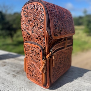 May include: A brown leather backpack with intricate floral tooling. The backpack features multiple zippered compartments and a top flap. The leather has a rich, warm tone, and the design is detailed with embossed flowers and leaves. The bag is set on a wooden surface.