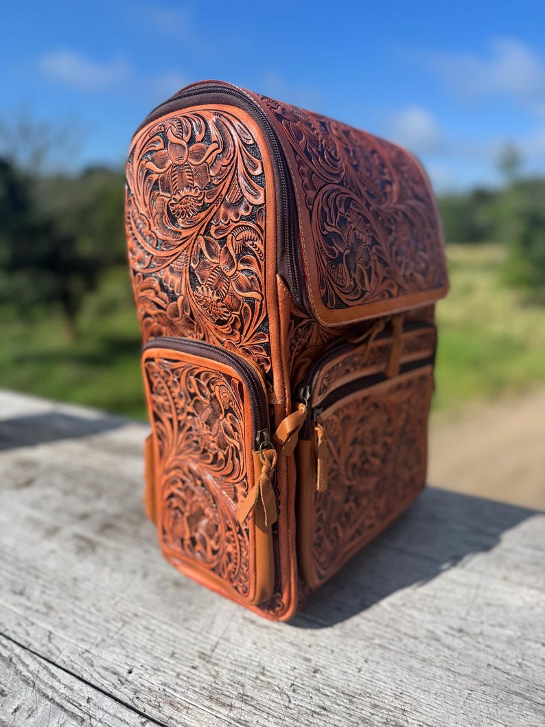May include: A brown leather backpack with intricate floral tooling. The backpack features multiple zippered compartments and a top flap. The leather has a rich, warm tone, and the design is detailed with embossed flowers and leaves. The bag is set on a wooden surface.