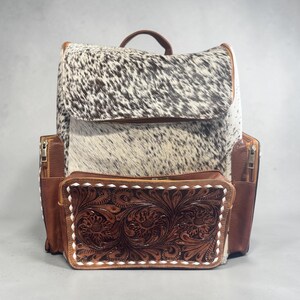 May include: A cowhide backpack with a brown leather top handle and trim. The front pocket features a detailed floral tooled leather design. The backpack has side pockets with zippers and a flap closure.