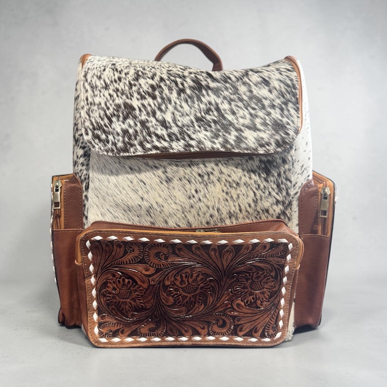 May include: A cowhide backpack with a brown leather top handle and trim. The front pocket features a detailed floral tooled leather design. The backpack has side pockets with zippers and a flap closure.