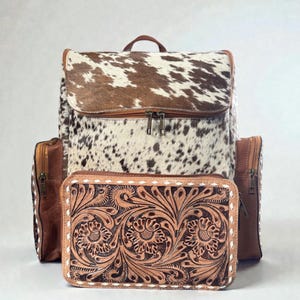 May include: A leather backpack with a cowhide flap and a matching wallet. The backpack has brown leather accents and a zipper closure. The wallet features a floral tooled design and white stitching.
