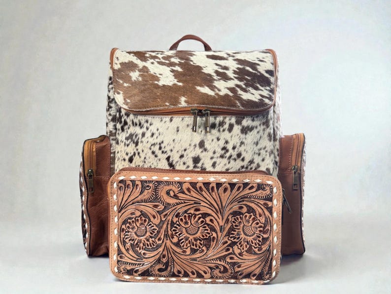 May include: A leather backpack with a cowhide flap and a matching wallet. The backpack has brown leather accents and a zipper closure. The wallet features a floral tooled design and white stitching.