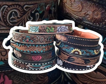 May include: A stack of intricately carved leather belts with colorful floral and geometric designs. The belts are brown, black, and turquoise with gold accents.