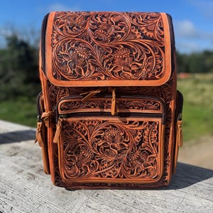 May include: A brown leather backpack with intricate floral carvings. The backpack features a flap closure, multiple zippered compartments, and adjustable straps. The detailed design includes embossed flowers and leaves, creating a rustic and stylish accessory.