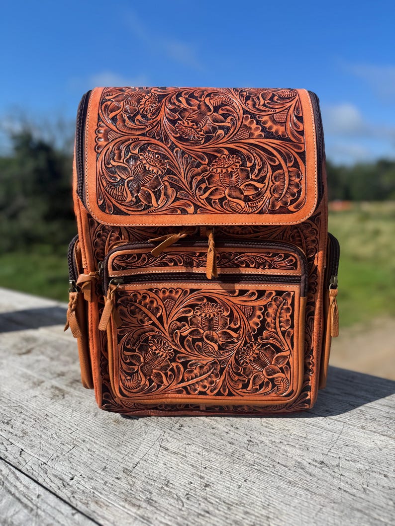May include: A brown leather backpack with intricate floral carvings. The backpack features a flap closure, multiple zippered compartments, and adjustable straps. The detailed design includes embossed flowers and leaves, creating a rustic and stylish accessory.