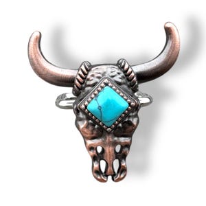 May include: A bronze-toned bull skull ring with curved horns and a turquoise-colored, diamond-shaped stone at the center. The ring has a detailed, textured design and is set against a white background.