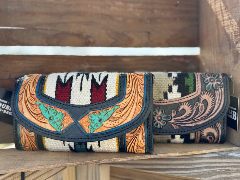 May include: Two leather wallets with tooled designs and colorful woven fabric accents. The wallets are closed and sitting on a wooden surface.