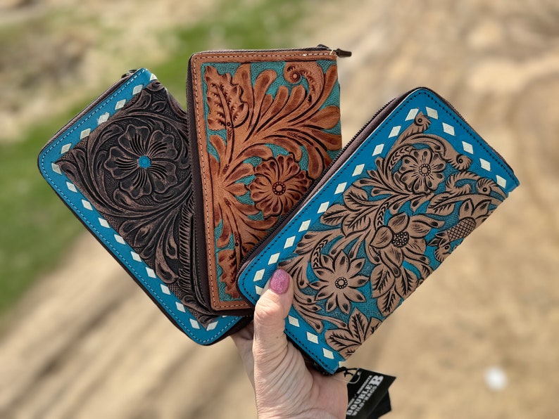 May include: Three leather wallets with floral tooling. The wallets feature a brown, turquoise, and white color scheme. Each wallet has a zipper closure and a blue border with white diamond accents. The wallets are held in a hand.