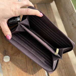 May include: Open brown zippered wallet with multiple card slots and compartments. The wallet has a gold-colored zipper and a small tag attached. The wallet is displayed on a wooden surface.