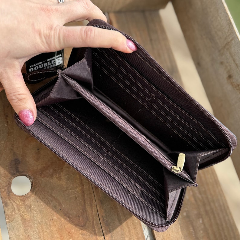 May include: Open brown zippered wallet with multiple card slots and compartments. The wallet has a gold-colored zipper and a small tag attached. The wallet is displayed on a wooden surface.