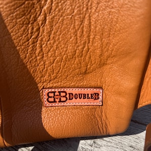 May include: Close-up of a textured, orange-brown leather item. A rectangular patch with the text "BB DOUBLER" in dark brown lettering is stitched onto the leather. The leather has a natural, slightly wrinkled appearance.