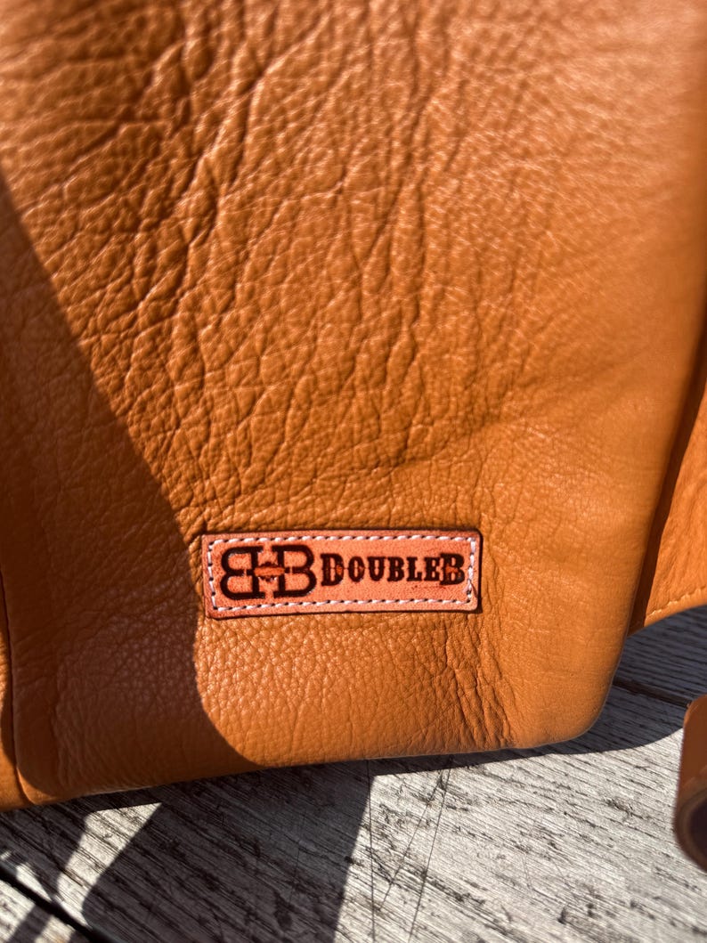 May include: Close-up of a textured, orange-brown leather item. A rectangular patch with the text "BB DOUBLER" in dark brown lettering is stitched onto the leather. The leather has a natural, slightly wrinkled appearance.