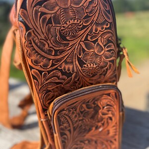 May include: A brown leather sling bag with intricate floral tooling. The bag features a main compartment with a zipper closure and a smaller front pocket. The leather has a rich, warm tone, and the floral design is deeply embossed.