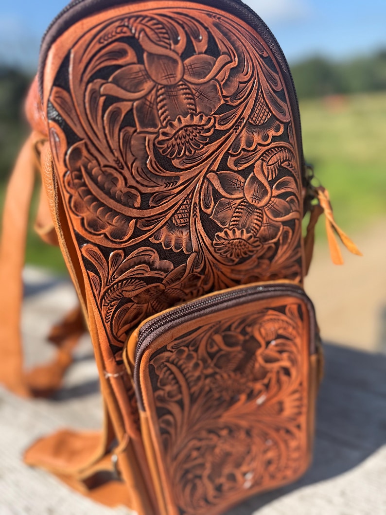 May include: A brown leather sling bag with intricate floral tooling. The bag features a main compartment with a zipper closure and a smaller front pocket. The leather has a rich, warm tone, and the floral design is deeply embossed.