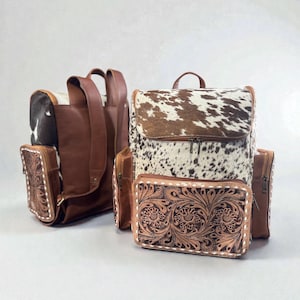May include: Two leather backpacks with a cowhide flap and a matching wallet. The backpacks are brown with a white and brown cowhide pattern on the flap. The wallet and side pockets have a floral tooled design.