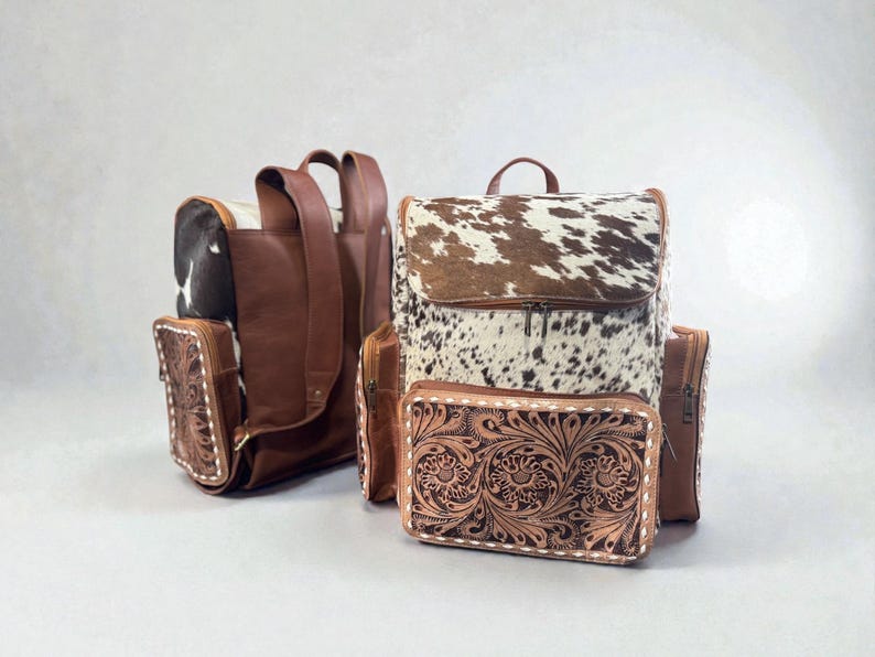 May include: Two leather backpacks with a cowhide flap and a matching wallet. The backpacks are brown with a white and brown cowhide pattern on the flap. The wallet and side pockets have a floral tooled design.