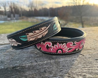 May include: Two black leather belts with intricate floral and feather designs. The belt on top has a turquoise and white feather design, while the belt on the bottom has a pink and white floral design.