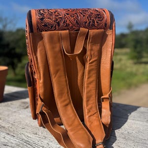 May include: A brown leather backpack with intricate floral tooling on the top flap. The backpack features adjustable shoulder straps and multiple pockets. The leather has a rich, warm tone, and the design suggests a handcrafted item.