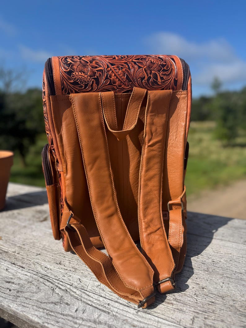 May include: A brown leather backpack with intricate floral tooling on the top flap. The backpack features adjustable shoulder straps and multiple pockets. The leather has a rich, warm tone, and the design suggests a handcrafted item.