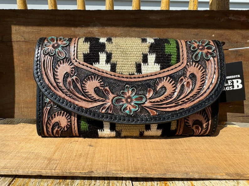 May include: A black leather wallet with a tooled floral design and a woven patterned flap. The wallet has a brown and green color scheme with turquoise accents. The wallet is labeled "Minnesota Saddle Bags".