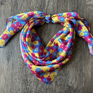 May include: A yellow, blue and pink floral patterned bandana tied in a knot. The bandana is made of a soft, silky fabric.
