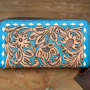 May include: A turquoise leather wallet with a floral design. The wallet features a brown zipper, a light brown floral pattern, and white diamond accents. The wallet is on a wooden surface.