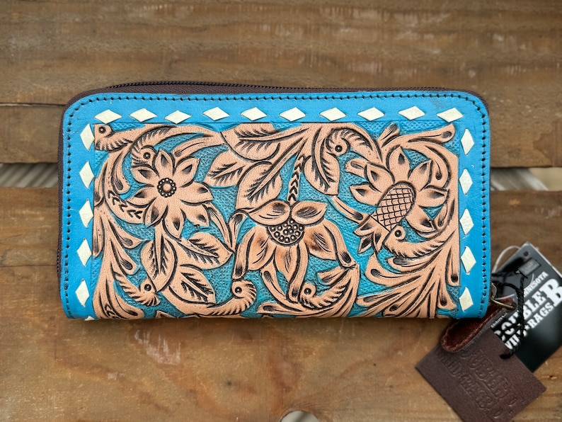 May include: A turquoise leather wallet with a floral design. The wallet features a brown zipper, a light brown floral pattern, and white diamond accents. The wallet is on a wooden surface.
