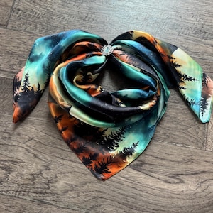 May include: A colorful silk scarf with a landscape design featuring trees, a night sky, and a decorative silver clasp. The scarf is primarily teal, orange, and black. The scarf is folded and arranged on a wooden surface.