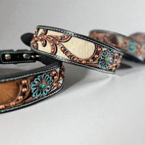 May include: Close-up of several decorative dog collars. The collars feature a black leather base with a light-colored fur center. They are adorned with copper-colored metal accents, turquoise-colored floral designs, and small copper studs.