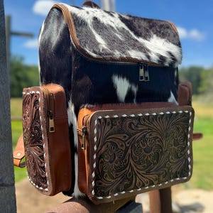 May include: A western-style backpack with a cowhide top and front flap, featuring a black and white pattern. The sides and pockets are brown leather with floral tooling and white stitching. The bag has a zipper closure and adjustable straps.