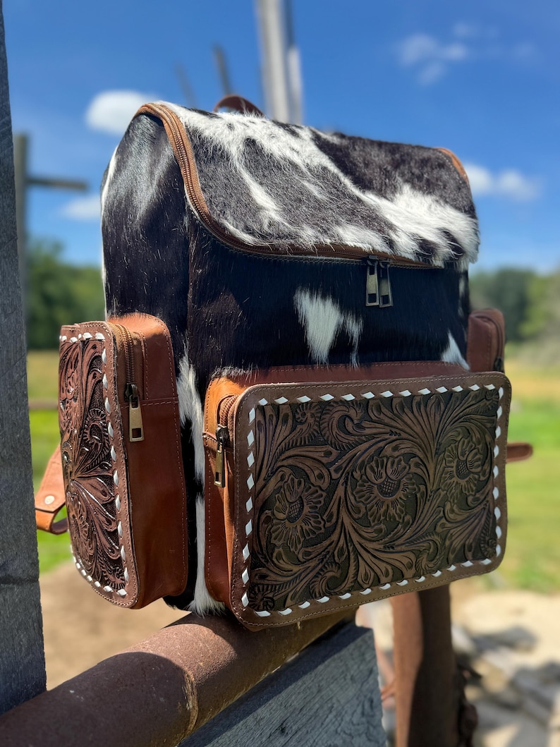 May include: A western-style backpack with a cowhide top and front flap, featuring a black and white pattern. The sides and pockets are brown leather with floral tooling and white stitching. The bag has a zipper closure and adjustable straps.