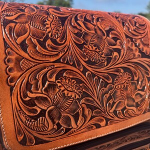 May include: Close-up of a brown leather backpack with intricate floral tooling. The embossed design features detailed flowers and swirling leaves, creating a textured surface. The bag has a zipper closure and a contrasting dark brown interior.