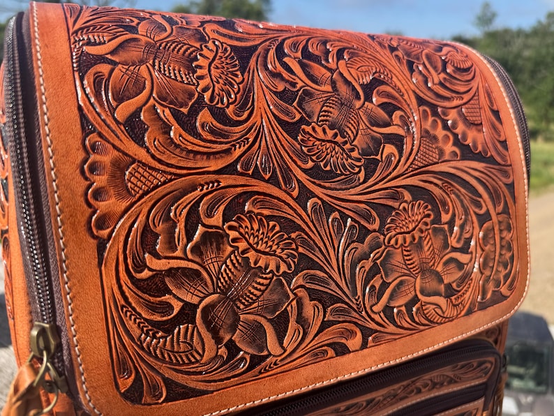 May include: Close-up of a brown leather backpack with intricate floral tooling. The embossed design features detailed flowers and swirling leaves, creating a textured surface. The bag has a zipper closure and a contrasting dark brown interior.