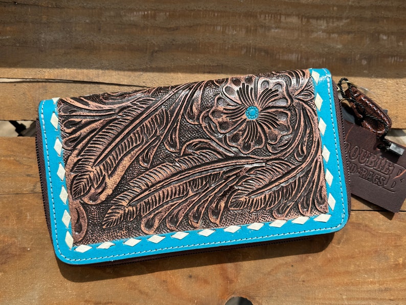 May include: A brown leather wallet with a floral and feather design. The wallet has a turquoise border with white stitching and a turquoise center stone in the flower. The wallet has a zipper closure and a tag that says "DOUBLE D RAGS D".