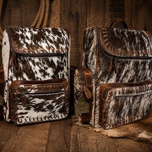 May include: Two cowhide backpacks with brown leather accents. The backpacks have a flap closure, front pocket, and side pockets. The cowhide pattern is brown and white. The backpacks are on a wooden surface.