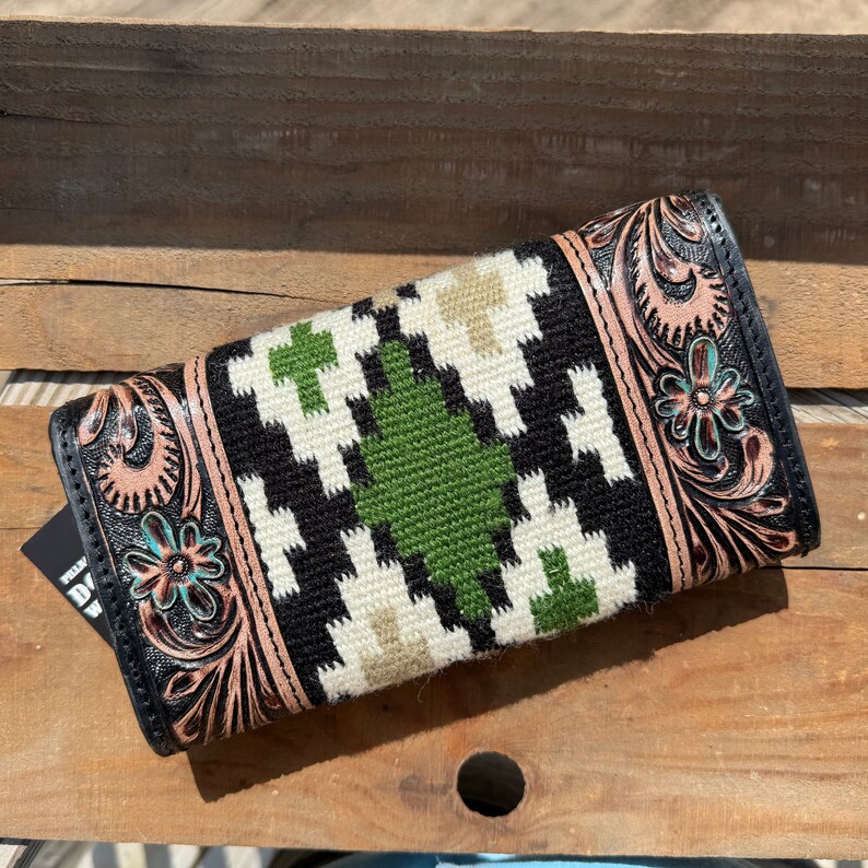 May include: A black leather wallet with a tooled floral design and a green, white, and black woven pattern. The wallet has a flap closure and a brown leather trim.